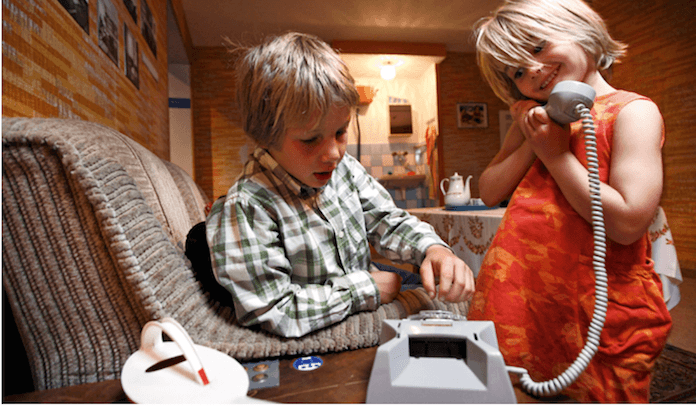 Spaß im Original-DDR-Wohnimmer. Das Telefon funktioniert. Die Plasteblumengießkanne steht für den Gummibaum bereit. Im Hintergrund das fensterlose Klo mit Deckenspülkasten und Wassermischbatterie mit Plastikdrehkappen für Kalt und Heiß (Foto: DDR Museum e.V. Berlin)